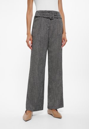 Frau trägt hochtaillierte, graue, weit geschnittene Hosen mit passendem Gürtel und beigefarbenen, spitz zulaufenden Ballerinas, stehend vor weißem Hintergrund.