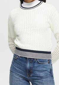 Weißer Strickpullover mit einem gerippten, marineblauen Saum und Kragen, der ein offenes Webmuster aufweist. Kombiniert mit blauen Jeans.