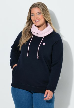 Smilende kvinde med langt hår iført en marineblå sweatshirt med en lyserød krave og jeans, stående mod en hvid væg.