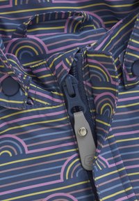 Blått tyg med flerfärgade regnbågs- och randmönster. Har en YKK-dragkedja med en ljusgrå dragknapp och tryckknapsstängningar.