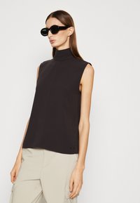 Top nero senza maniche con scollatura alta, texture liscia e design minimalista, abbinato a occhiali da sole ovali scuri.