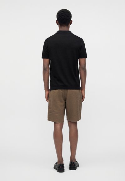 Homme debout, dos tourné, portant un polo noir à manches courtes, un short marron et des chaussures noires sur un fond blanc uni.
