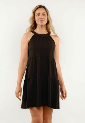 Žena s dlhými vlnitými vlasmi, oblečená v čiernych bezrukávoch s halterovým výstrihom, stojaca pred jednoduchým svetlým pozadím.