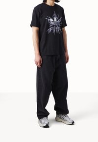 Black cotton t-shirt featuring a white graphic of three stripes and lightning pattern. Paired with loose black denim jeans and grey shoes.