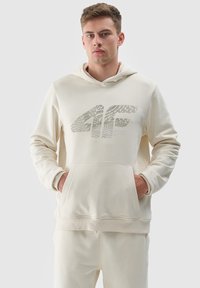 Crème-kleurige hoodie van zacht materiaal, met een voorzak in kangoeroestijl en een grafisch logo ontwerp op de borst.