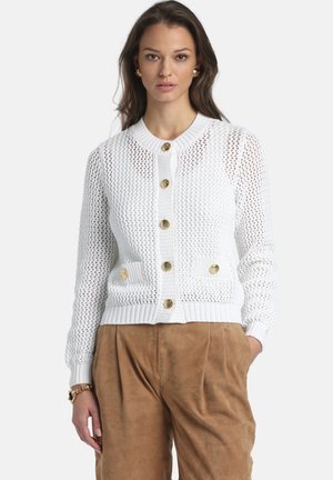 Vrouw draagt een witte gebreide cardigan met een structuur en gouden knopen, en lichtbruine suède broek, staand tegen een effen achtergrond.