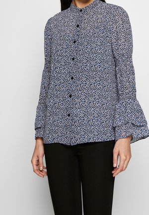Femme portant un chemisier bleu et noir fleuri à manches longues avec des poignets à volants et un pantalon noir sur un fond uni.