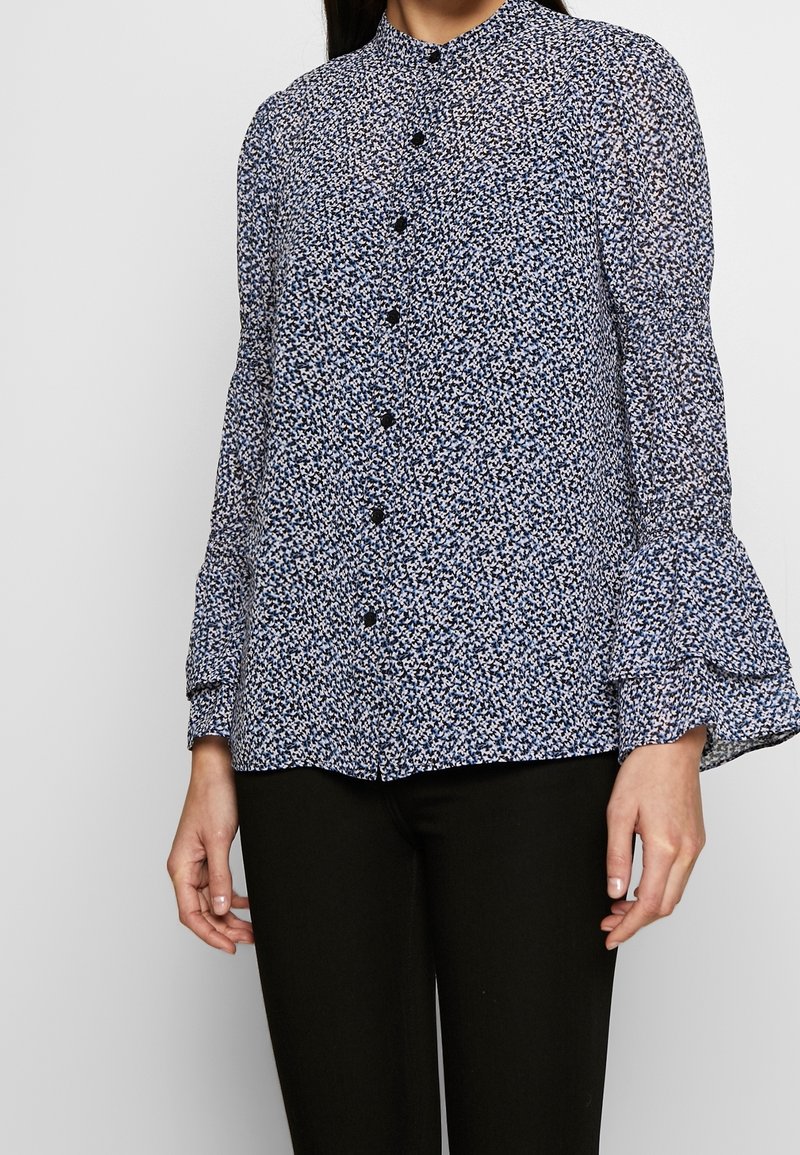 Femme portant un chemisier bleu et noir fleuri à manches longues avec des poignets à volants et un pantalon noir sur un fond uni.