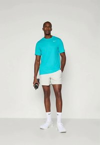 Hombre con camiseta turquesa de Nike, pantalones cortos blancos, calcetas blancas de Nike y zapatillas blancas, sosteniendo una botella de agua negra, de pie en un espacio neutral.