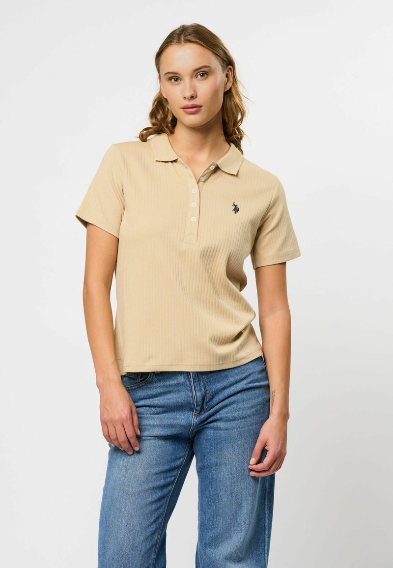 Beige geribbelde polo met korte mouwen, drie knopen aan de voorkant en geborduurd logo op de borst, gedragen met lichtblauwe jeans.