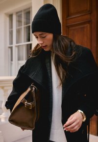 Bonnet côtelé noir, manteau noir à larges revers, couche intérieure grise, pull blanc, tenant un sac à main en daim marron avec des accents métalliques.