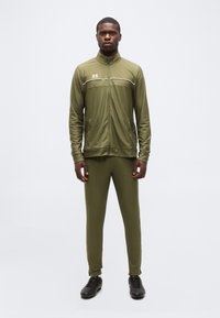 Homme debout devant un fond uni, portant une veste de survêtement à fermeture éclair intégrale olive et un pantalon assorti, avec des chaussures de sport noires.