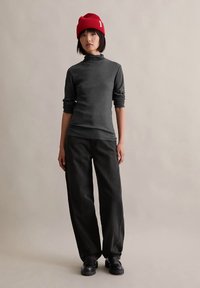 Jovem a usar um gorro vermelho, um pulôver de gola alta cinza escuro, calças largas pretas e sapatos pretos, de pé contra um fundo beige liso.