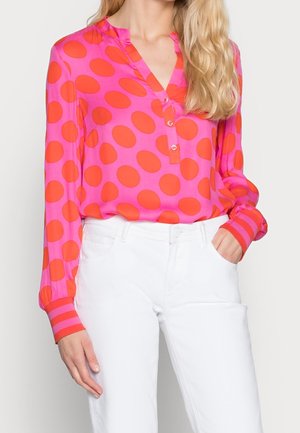 Kvinde iført en pink bluse med store røde polkaprikker og stribede manchetter, kombineret med højtaljede hvide bukser, hånd i lommen.