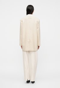 Ανοιχτό εκρού κοστούμι με μακρύ σακάκι και παντελόνες φαρδιάς γραμμής. Το ύφασμα φαίνεται υφασμένο, με κομψό, μίνιμαλ σχεδιασμό και λεπτομέρειες με ανοίγματα στο πίσω μέρος.