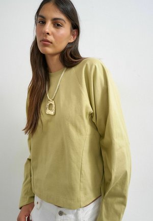 Jonge vrouw met lang bruin haar die een lichtgroene trui, witte broek en een grote gouden hanger aan een touwketting draagt.