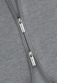 Szara kurtka zapinana na zamek wykonana z miękkiego materiału, z metalowymi suwakami z akcentami logo oraz gładką fakturą.