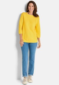 Gelbes Strickoberteil mit Dreiviertelärmeln, kombiniert mit hellblauen Jeans, die an den Bündchen hochgekrempelt sind, und beigen Sneakers.