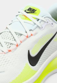 Nike trkaće tenisice u bijeloj boji s neonsko žutim detaljima. Imaju prozračnu mrežastu gornju površinu, crni logo i teksturirani potplat. Svijetle vezice upotpunjuju dizajn.