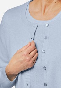 Hellblauer Cardigan aus weichem Material, mit rundem Hals, Knopfverschluss und gerippten Bündchen. Die Knöpfe sind in einem passenden Farbton.
