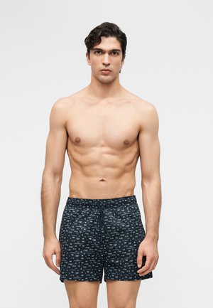 Νέος άνδρας με σκούρα μαλλιά που στέκεται, φοράει μαύρο μαγιό με σχέδια, μπροστά σε απλό φωτεινό φόντο.