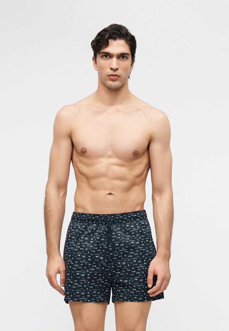 Νέος άνδρας με σκούρα μαλλιά που στέκεται, φοράει μαύρο μαγιό με σχέδια, μπροστά σε απλό φωτεινό φόντο.