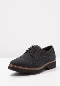 Chaussure noire en suede à lacets avec des détails brogues, bout arrondi, coutures brunes et semelle en caoutchouc texturé pour une meilleure adhérence et stabilité.