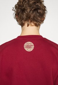 Felpa bordeaux realizzata in tessuto morbido, presenta un logo tondo Carhartt beige con testo, posizionato sulla parte superiore della schiena.
