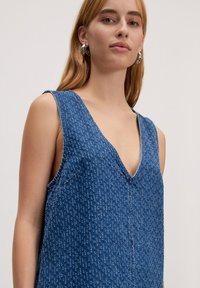 Denim jurk in donkerblauw, met een diepe V-hals en brede schouderbanden. De stof heeft een subtiel textuurpatroon doorheen.