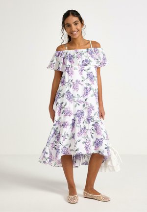 Ung kvinde smilende, iført en hvid off-shoulder kjole med lilla blomsterprint og beige ballet flats, stående foran en ensfarvet baggrund.