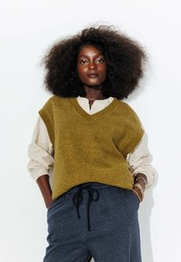 Gilet texturé vert olive avec un col en V porté par-dessus une chemise à rayures à boutons, associé à un pantalon à carreaux foncé avec cordon de serrage.