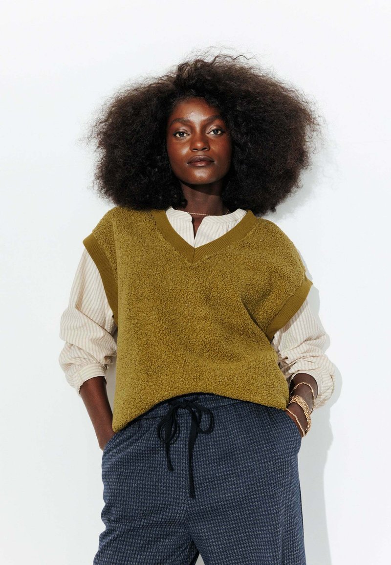 Gilet texturé vert olive avec un col en V porté par-dessus une chemise à rayures à boutons, associé à un pantalon à carreaux foncé avec cordon de serrage.