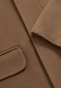 Brauner Wollmantel im Nahaufnahme, mit weicher Textur, Reverskragen und aufgesetzter Tasche. Der Stoff wirkt dick und warm.