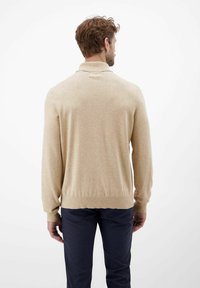 Beiges Strickpullover mit geripptem Kragen und Bündchen, taillierte Passform und glatter Textur, über einer dunkelblauen Hose getragen, von hinten betrachtet.