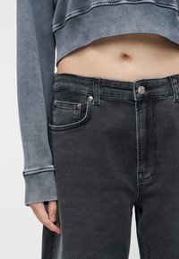 Μέσος κορμός ατόμου που φοράει γκρι κοντή μπλούζα και ψηλόμεσα ξεθωριασμένα μαύρα τζιν, με το χέρι ακουμπισμένο στο πλευρό.