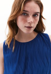 Abito blu a pieghe con scollatura rotonda. Tessuto liscio con texture a onde verticali. La modella ha capelli castano chiaro e lentiggini.