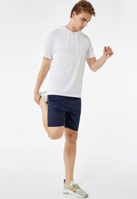 Valkoinen lyhythihainen huppari, jossa on huppu, yhdistettynä tummansinisiin urheilusortseihin. Malli venyttelee, jalassa vaaleansävyiset urheilukengät.