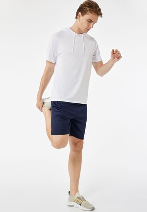 Valkoinen lyhythihainen huppari, jossa on huppu, yhdistettynä tummansinisiin urheilusortseihin. Malli venyttelee, jalassa vaaleansävyiset urheilukengät.