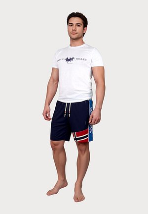 Muž stojaci bosý, oblečený v bielom tričku s logom Harvey Miller a tmavomodrých šortkách s červenými, bielymi a modrými vzorovanými detailmi.