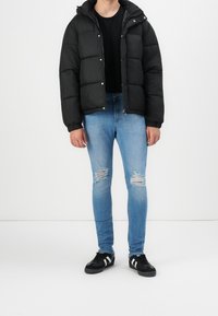 Chaqueta de plumón negra con capucha, cierre frontal y botones a presión; combinada con jeans desgastados de color azul claro y zapatillas negras con rayas blancas.