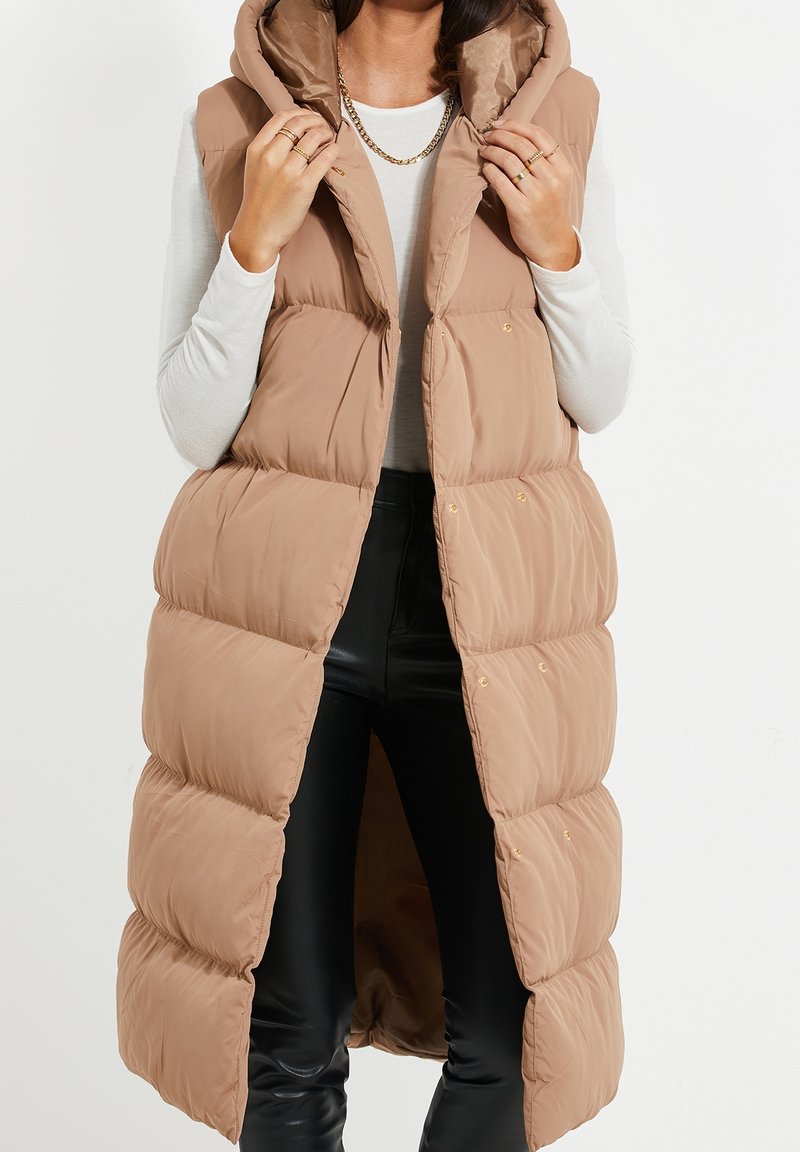 Femme portant une longue doudoune beige sur un haut blanc à manches longues et un pantalon en cuir noir, tenant le col de la doudoune avec les deux mains.