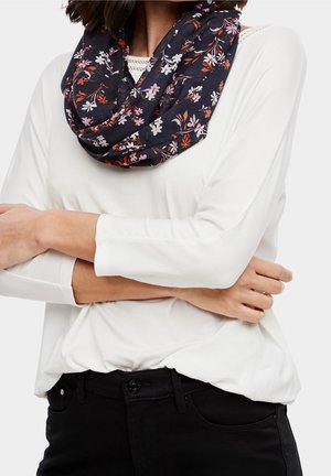 Personne portant une chemise blanche à manches longues, un pantalon noir et une écharpe infinie à motifs floraux foncés avec des fleurs roses et orange.