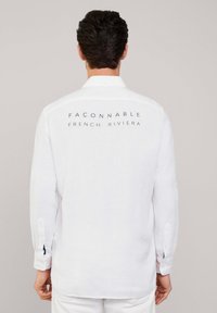 Camisa blanca de lino de manga larga con cuello abotonado, que presenta el texto "FAÇONNABLE FRENCH RIVIERA" en azul marino en la parte posterior.
