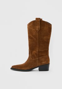 Καφέ σουέτ δυτικό μποτάκι μεσαίου μήκους μέχρι τη γάμπα, με μυτερή μύτη, θηλιές για κράτημα και ψηλή μαύρη σόλα, πάνω σε ανοιχτό γκρι φόντο.