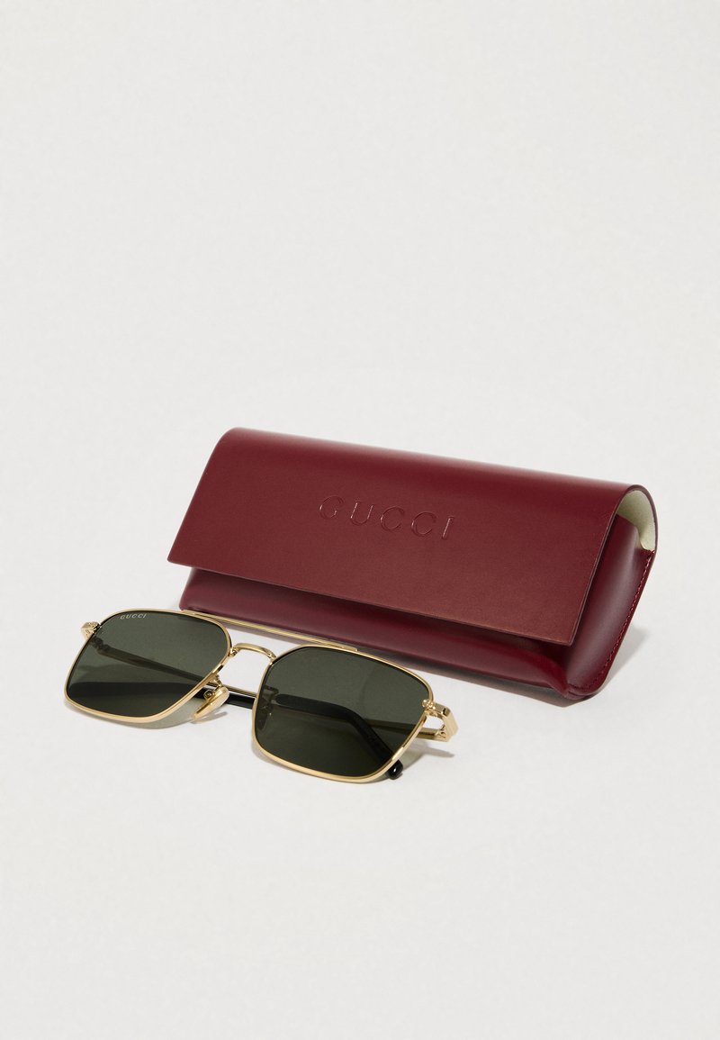 Gucci zonnebril met een gouden metalen frame, donkere groene lenzen en zwarte pootjes, vergezeld van een bordeauxleren etui met het opschrift "Gucci".