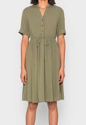 Femme portant une robe olive verte longueur genou avec manches courtes, boutonnée à l'avant, et ceinture à la taille, debout devant un fond clair uni.