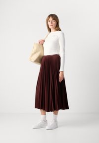 Jupe plissée brun, haut à manches longues blanc, baskets blanches, tenant un sac fourre-tout en toile beige à motifs. Texture lisse, coupe décontractée.