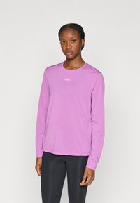 Ελαφρύ πουκάμισο μακρυμάνικο σε έντονο ροζ, με στρογγυλή λαιμόκοψη και διακριτή υφή· το λογότυπο είναι τοποθετημένο στο κέντρο του στήθους. Συνδυασμένο με μαύρα κολάν.