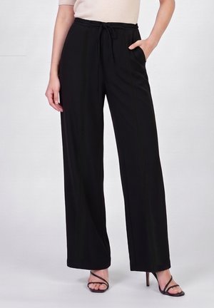 Femme portant un pantalon noir taille haute à jambes larges avec cordon à la taille et poches latérales, associé à un haut beige et des talons noirs à brides.