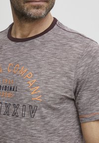 Short-sleeve, crew neck t-shirt in heathered burgundy with contrasting maroon collar. Features textured fabric and printed logo in orange and black.